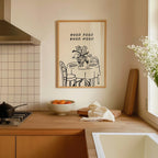 a kitchen counter with a bowl of fruit, a vase of flowers, and a framed poster with a table and chairs.