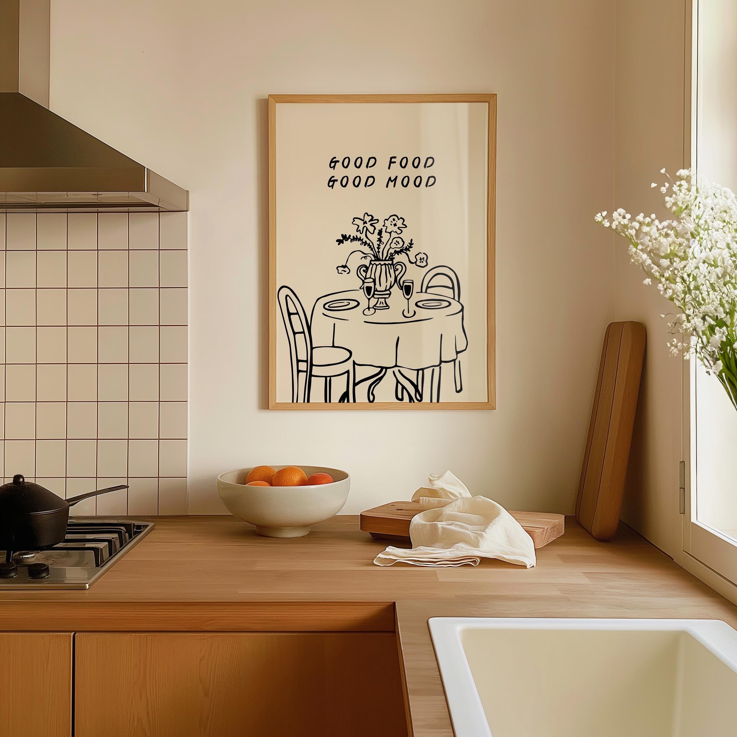 a kitchen counter with a bowl of fruit, a vase of flowers, and a framed poster with a table and chairs.