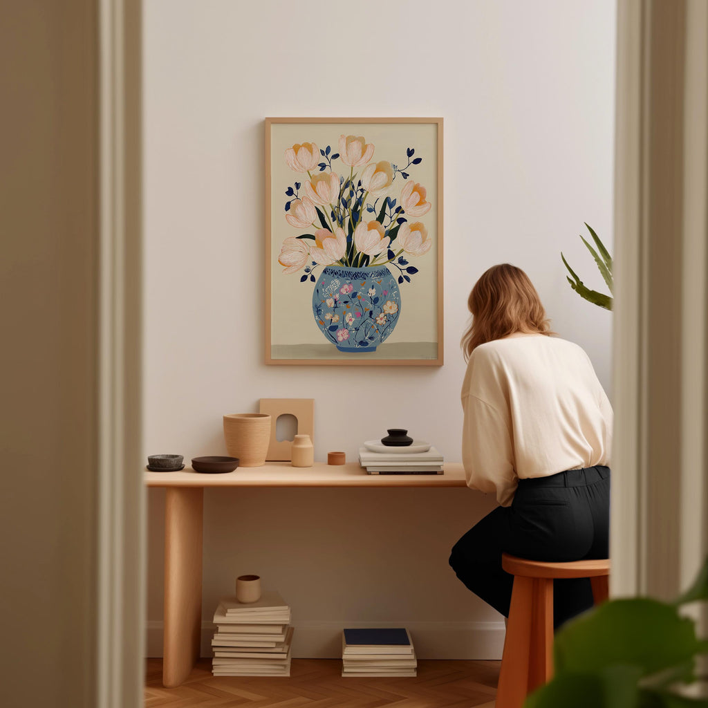 A woman is sitting at a wooden desk, looking at a framed painting of flowers on the wall. The desk has various items on it, including a vase, a stack of books, and a plant.