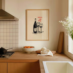 a kitchen counter with a framed drawing of a wine bottle and two wine glasses, along with a bowl of fruit and a vase of flowers.