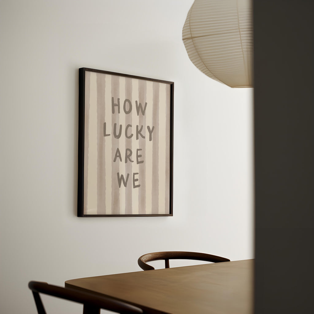 a framed poster with the text "HOW LUCKY ARE WE" hanging on a wall above a wooden table with chairs.