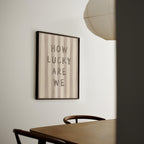 a framed poster with the text "HOW LUCKY ARE WE" hanging on a wall above a wooden table with chairs.