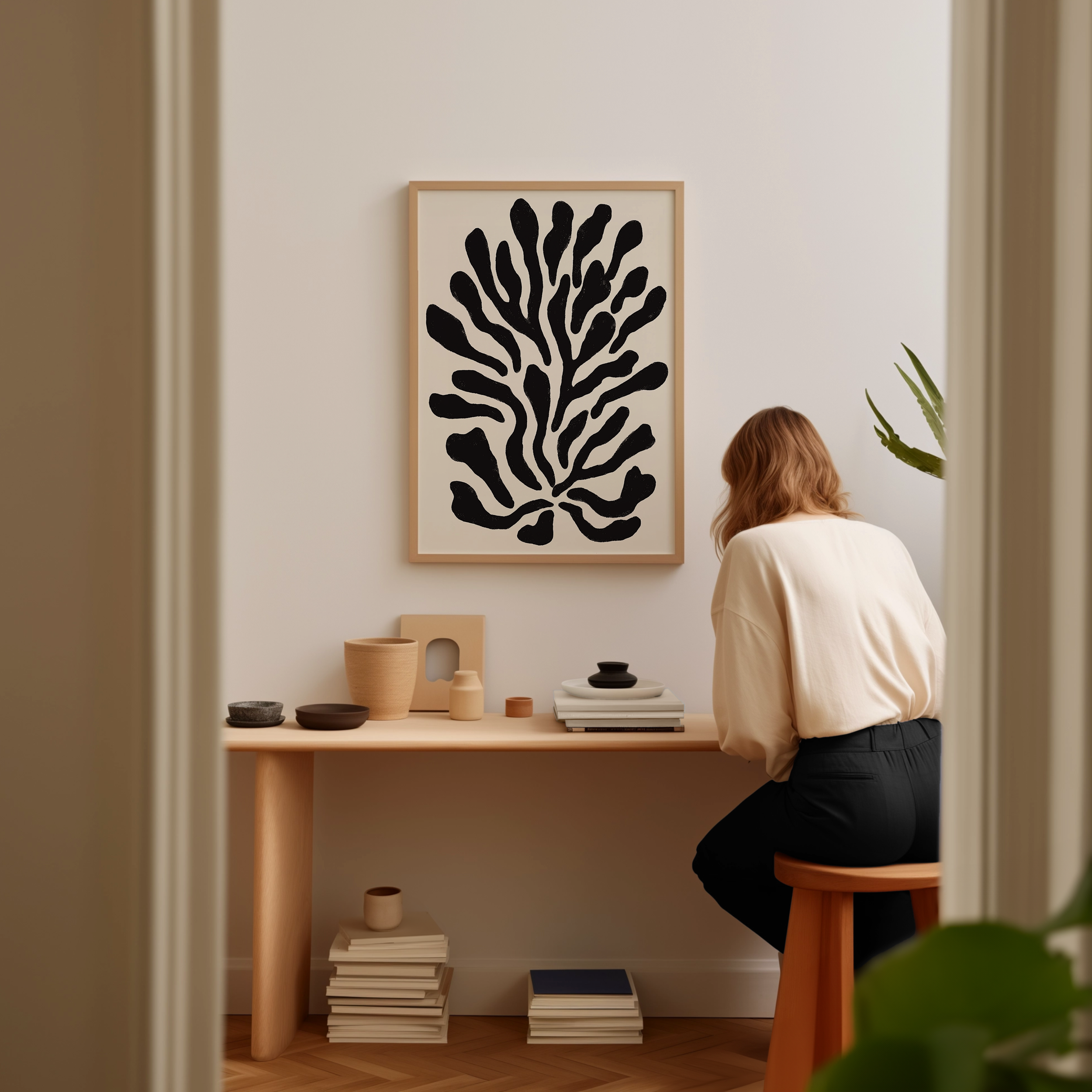 A woman is sitting at a wooden desk, surrounded by various objects, including a plant, books, and a framed abstract art piece on the wall.