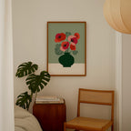 a cozy corner of a room with a framed artwork of a vase with red flowers on the wall, a wooden chair, a stack of books, and a potted plant.