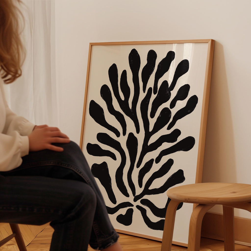 A person is sitting on a chair in front of a large framed artwork depicting a stylized black and white plant or flower design.