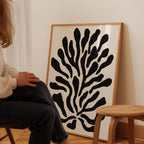 A person is sitting on a chair in front of a large framed artwork depicting a stylized black and white plant or flower design.
