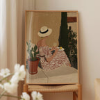 A framed painting depicting a woman in a striped dress sitting on a bench, holding a glass of wine and surrounded by potted plants.