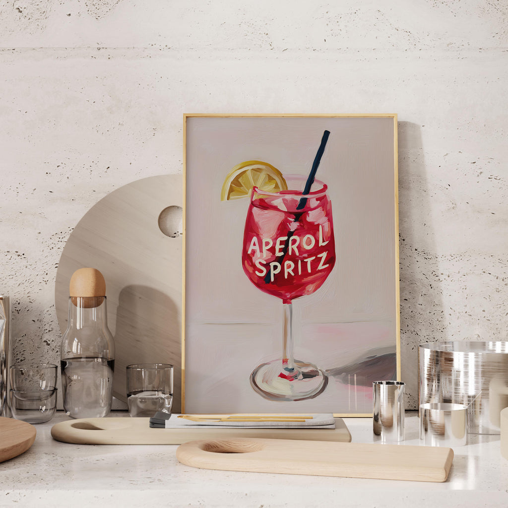 a framed painting of a red drink with a lemon slice and the text "Aperol Spritz" on it, placed on a table with various kitchen items such as a bottle, cups, and a cutting board.
