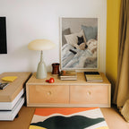 a cozy corner of a room with a wooden dresser, a lamp, and a framed picture of a cat sleeping on a bed. The dresser has two drawers and a small table with a lamp and a red object on it. The framed picture adds a personal touch to the space.