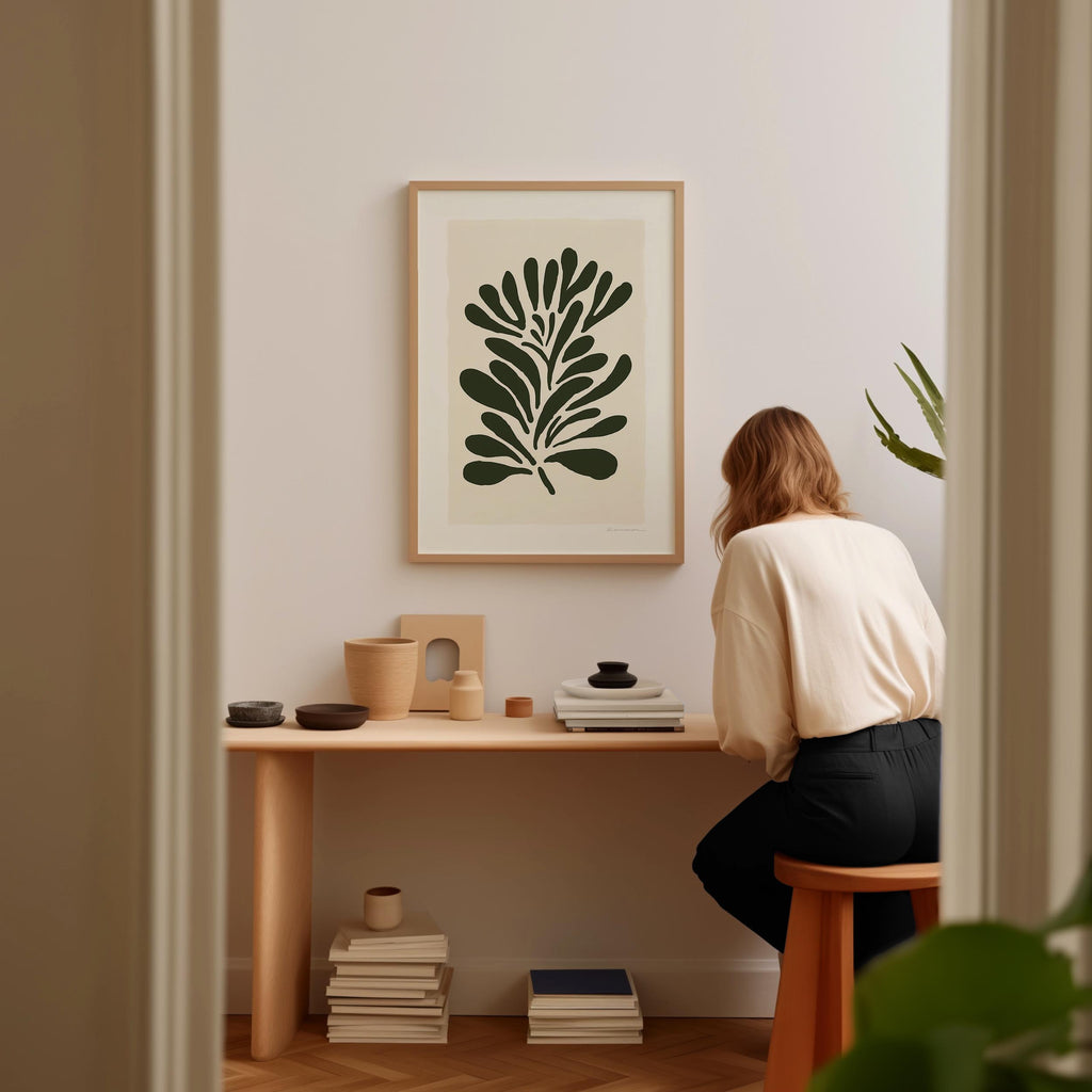 A woman is sitting at a wooden desk, surrounded by various objects, including a plant, books, and a framed artwork on the wall.