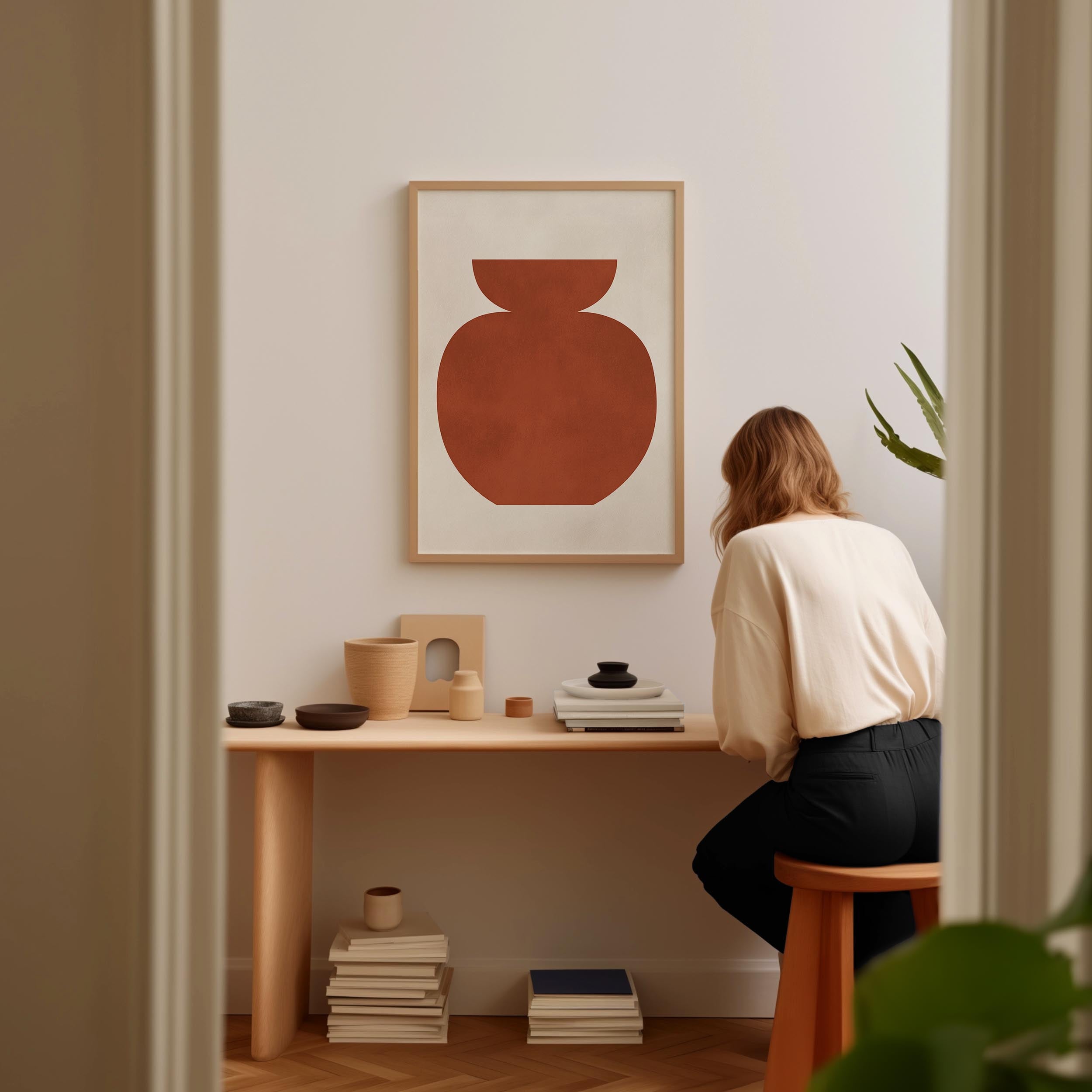 A woman is sitting at a wooden desk, surrounded by various objects, including a vase, books, and a plant, in a minimalist and modern interior setting.