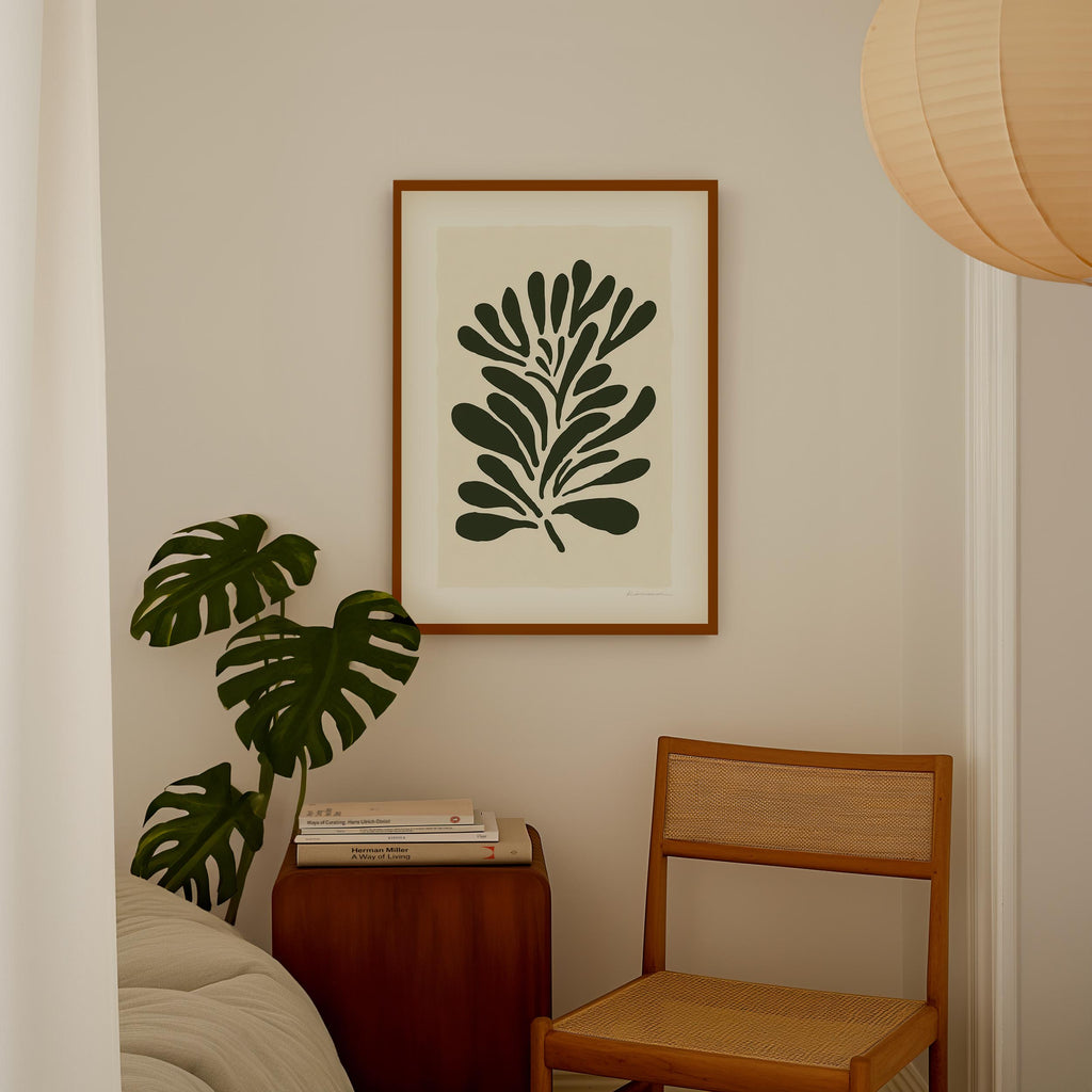a cozy corner of a room with a framed artwork of a leafy plant on the wall, a wooden chair, and a stack of books on a wooden table.